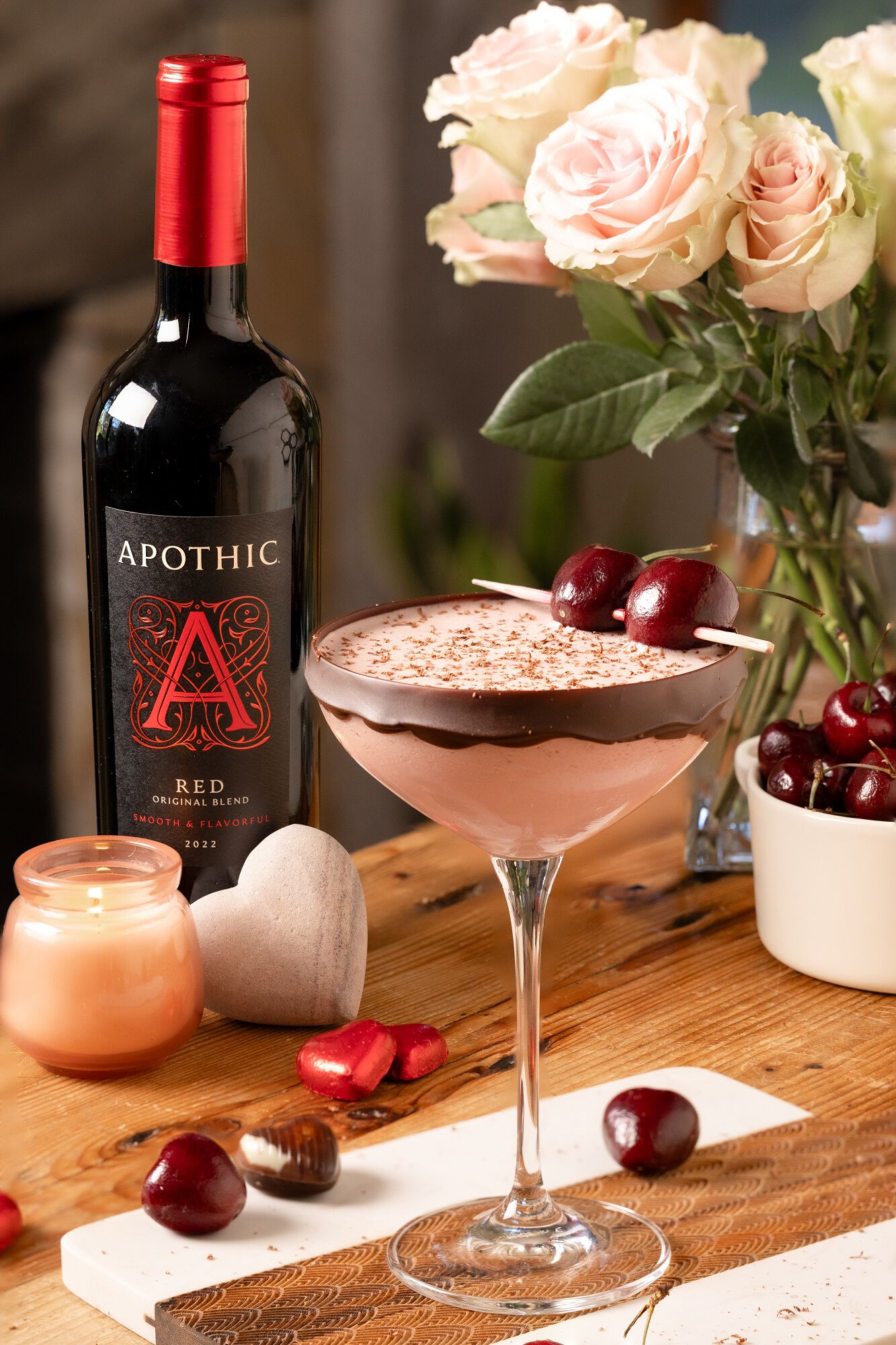 A sophisticated lifestyle shot of a Chocolatini served in a chilled martini glass on a dark, reflective surface. The deep red drink is topped with a dense layer of creamy foam and garnished with a single maraschino cherry. In the background, a bottle of Apothic Red Original Blend stands next to a bowl of fresh cherries and pieces of dark chocolate. The lighting is focused and moody, creating elegant reflections on the glassware.