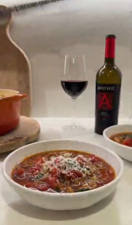A warm lifestyle photograph featuring a white ceramic bowl of hearty sausage and pasta soup topped with shredded parmesan cheese and fresh herbs. In the background, a bottle of Apothic Red Original Blend and a filled wine glass sit on a white countertop. An orange Dutch oven on a wooden board is partially visible to the left, creating a cozy and inviting home-cooked meal scene.
