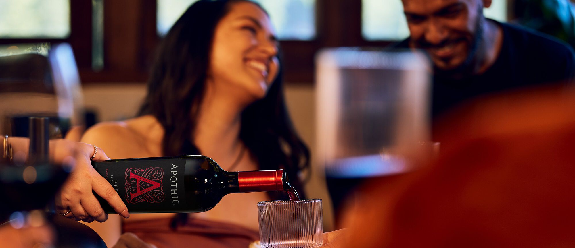 A warm, lifestyle banner image showing a group of friends laughing and socializing in a dimly lit setting. In the foreground, a person's hand pours Apothic Red Original Blend from a black bottle with a vibrant red 'A' logo into a modern, ribbed glass. The background is softly blurred, focusing the attention on the act of sharing the wine.