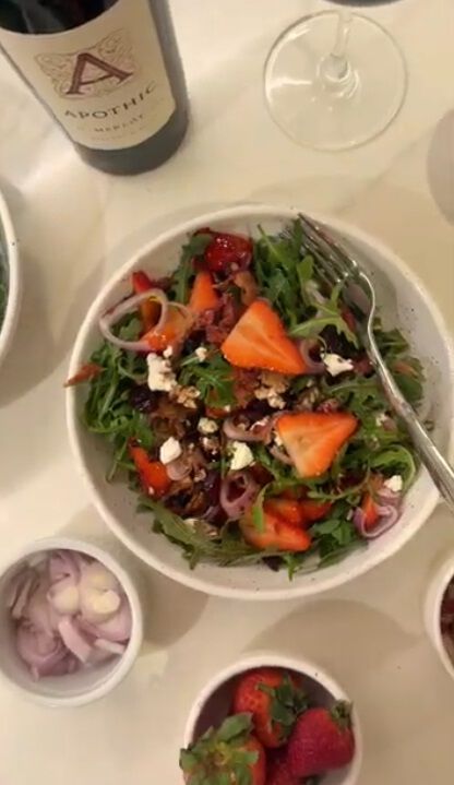 A top-down, close-up shot of a fresh arugula and berry salad in a white ceramic bowl. The salad features green arugula leaves topped with sliced strawberries, red onion slivers, crumbled feta cheese, and walnuts. In the upper left corner, the corner of an Apothic Merlot wine bottle and a glass of red wine are visible on a white tabletop, alongside small side bowls of sliced onions and whole strawberries.