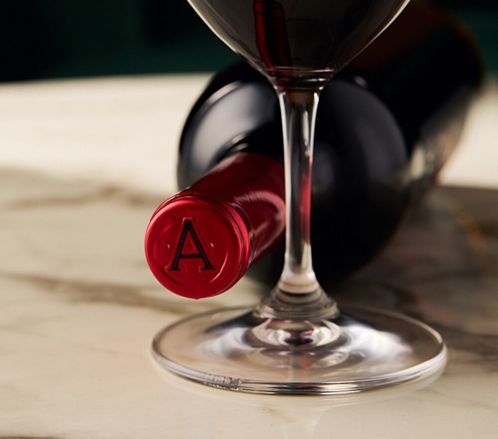 A close-up shot focusing on the red cap of an Apothic Red wine bottle lying down, which is marked with a raised letter 'A'. The stem of a wine glass partially filled with red wine stands in the foreground, with the bottle extending into the background on a marble surface.