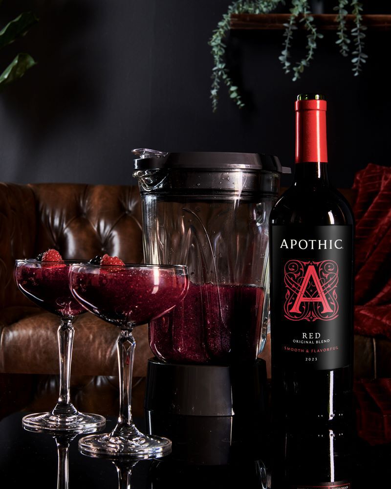 A moody lifestyle shot of two stemmed coupe glasses filled with a deep purple frozen Red Slush cocktail, garnished with fresh raspberries and blackberries. A blender containing the remaining blended wine and fruit mixture sits behind the glasses. To the right, a bottle of 2023 Apothic Red Original Blend stands on a dark, reflective surface. The background features a tufted brown leather sofa and a dark wall with a hint of green foliage.