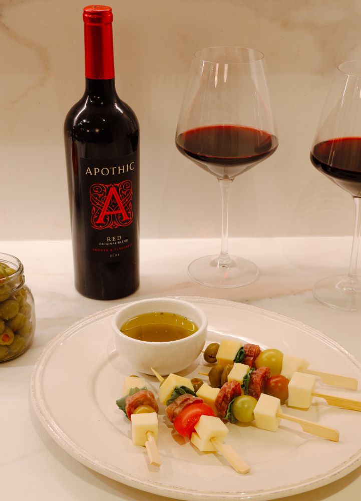A bright, overhead shot of a white plate holding five antipasto skewers with cheese, olives, meat, and tomatoes, served with a small bowl of dipping oil. In the background, a bottle of 2022 Apothic Red Original Blend stands next to two glasses filled with red wine on a white marble surface. A jar of green olives is visible on the left, creating a fresh and inviting appetizer scene.