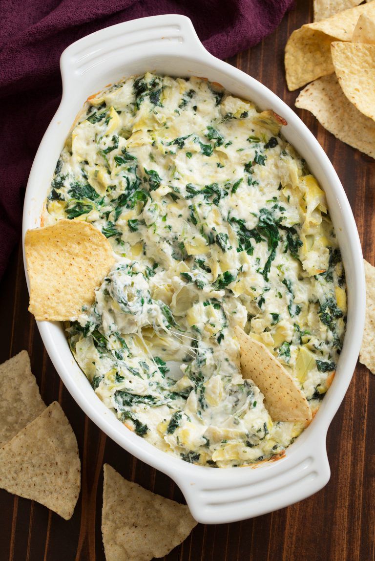 A top-down lifestyle shot of a warm, bubbly spinach and artichoke dip served in an oval white ceramic baking dish. Tortilla chips are scattered around the dish on a dark wooden tabletop, with one chip scooping up a portion of the melted, cheesy dip. A maroon cloth napkin is visible in the corner, complementing the rich tones of the culinary scene.