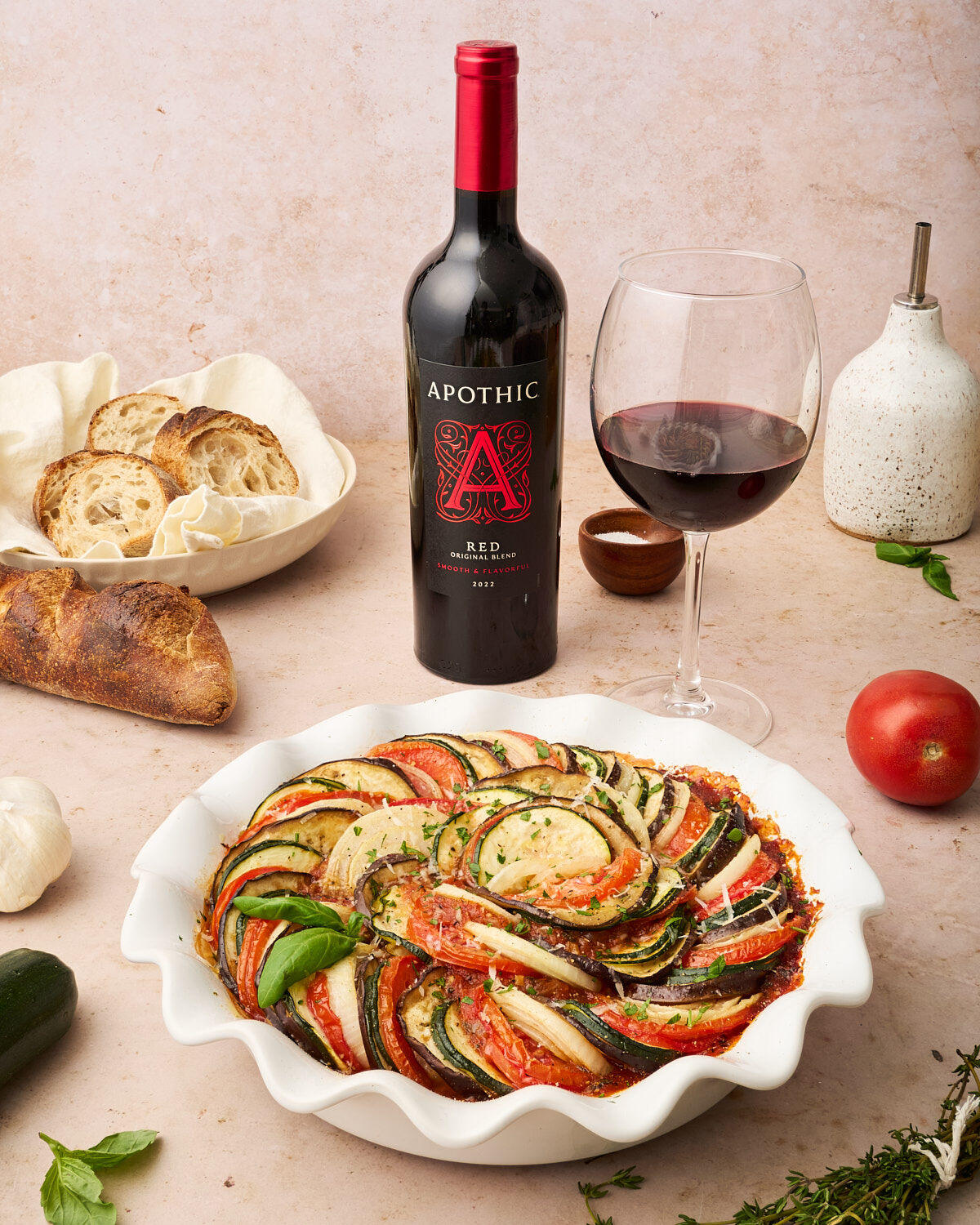 A bottle of Apothic Red 2022 wine and a glass of red wine are served alongside a colorful, fresh vegetable ratatouille in a white ruffled baking dish. The scene is set on a light-textured surface with rustic bread, a whole tomato, garlic, and fresh herbs scattered around.