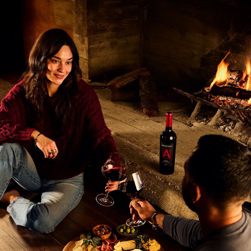 A warm, atmospheric lifestyle shot of a couple enjoying an indoor date night by a glowing fireplace. A woman in a maroon sweater sits on the floor across from her partner, both holding large glasses of red wine. Between them on a low stone ledge is a bottle of Apothic Red Original Blend. In the foreground, a wooden charcuterie board is spread with olives, nuts, and cheeses. The low lighting from the fire creates a cozy, private VIP experience at home.