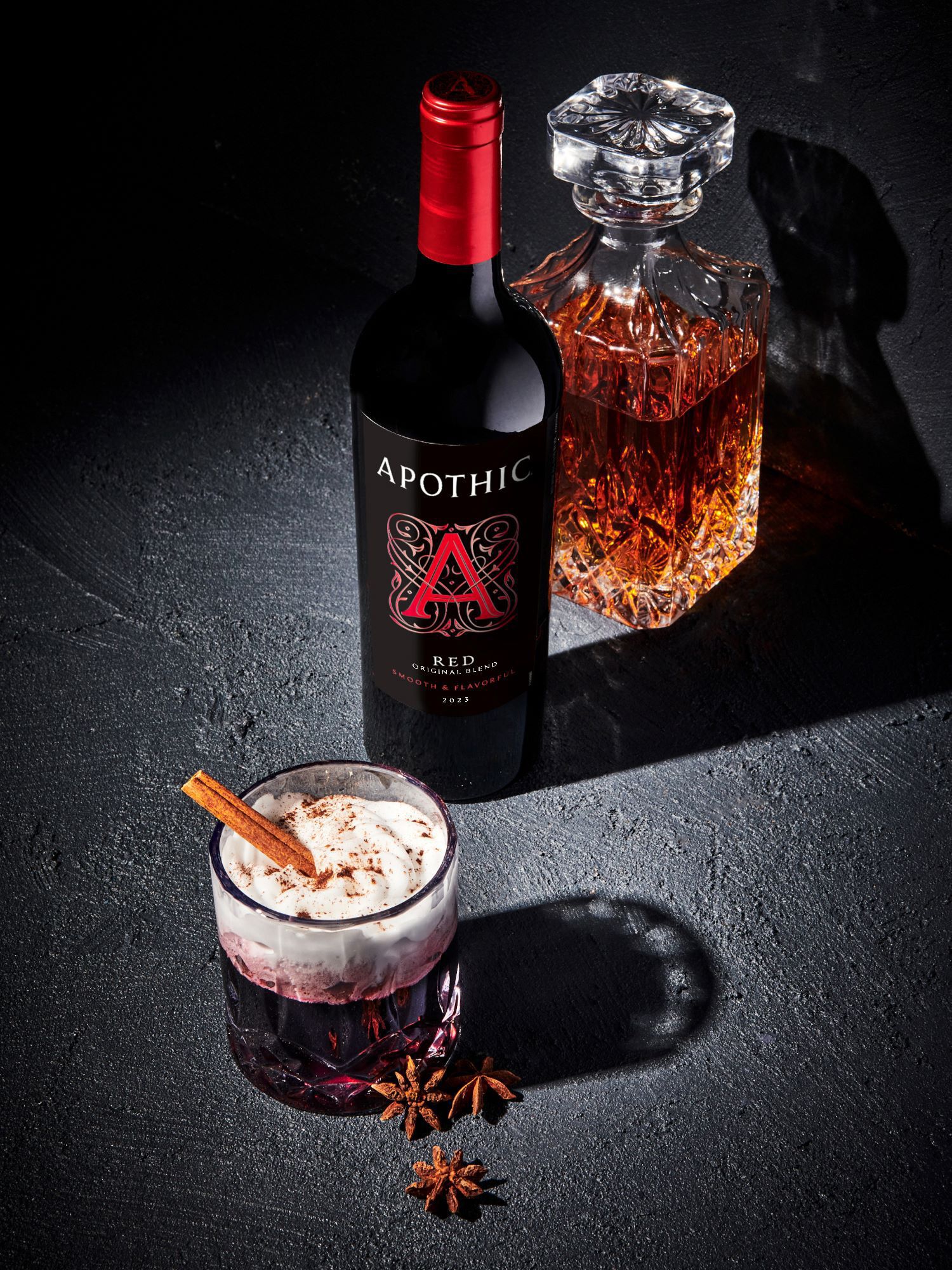 A moody, high-contrast photograph of a winter wine cocktail on a dark textured surface. In the foreground, a crystal rocks glass is filled with a dark red drink topped with a thick layer of whipped cream, dusted with cocoa and garnished with two cinnamon sticks. Next to the glass lie two whole star anise. Behind the cocktail stands a bottle of 2023 Apothic Red Original Blend and a crystal decanter filled with amber-colored bourbon.