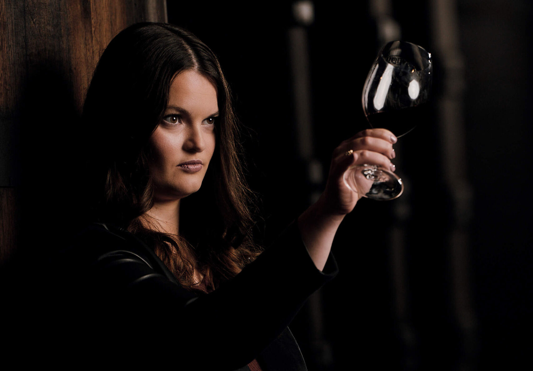A moody, low-light portrait of Apothic winemaker Ashleigh 'Ash' Ricchio. She is leaning against a dark wooden surface, wearing a black leather jacket, and looking thoughtfully off-camera. She holds a glass of deep red wine up to the light, which catches the rim of the glass and highlights her face against the dark, shadowed background.