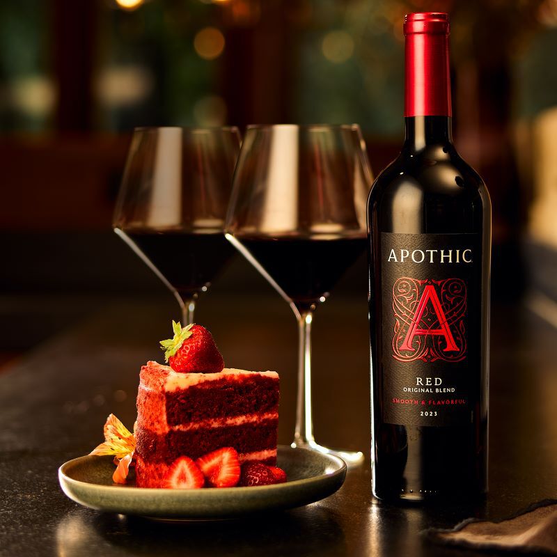 A close-up lifestyle photograph of a decadent date night pairing. A slice of rich red velvet cake topped with fresh strawberries sits on a small plate next to a bottle of 2023 Apothic Red Original Blend. Two filled wine glasses stand in the softly blurred background on a dark tabletop. The warm, amber lighting emphasizes the deep colors of the wine and the indulgent texture of the dessert.