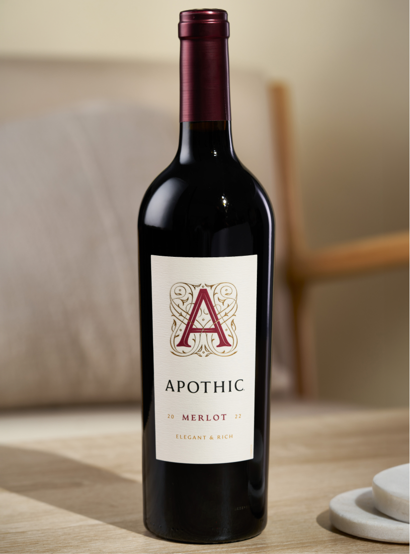A centered, eye-level photograph of a bottle of 2022 Apothic Merlot. The bottle features a textured white label with a maroon 'A' logo bordered in gold and the descriptors 'Elegant & Rich.' It is set on a light-colored wooden surface in a bright, modern interior with a soft-focus cream armchair in the background.