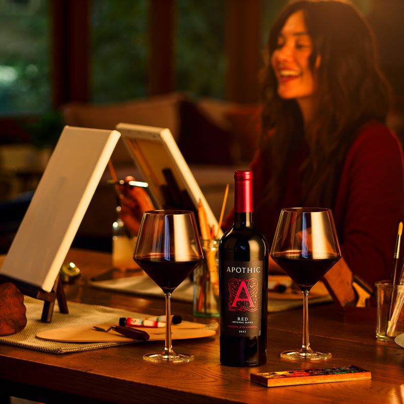 A warm, atmospheric lifestyle photograph of a couple enjoying a creative date night at home. In the center, a bottle of 2023 Apothic Red Original Blend sits between two filled wine glasses on a wooden table. In the blurred background, a woman smiles while painting on a canvas. Art supplies, including brushes and a palette, are scattered across the table, creating a cozy and artistic scene.