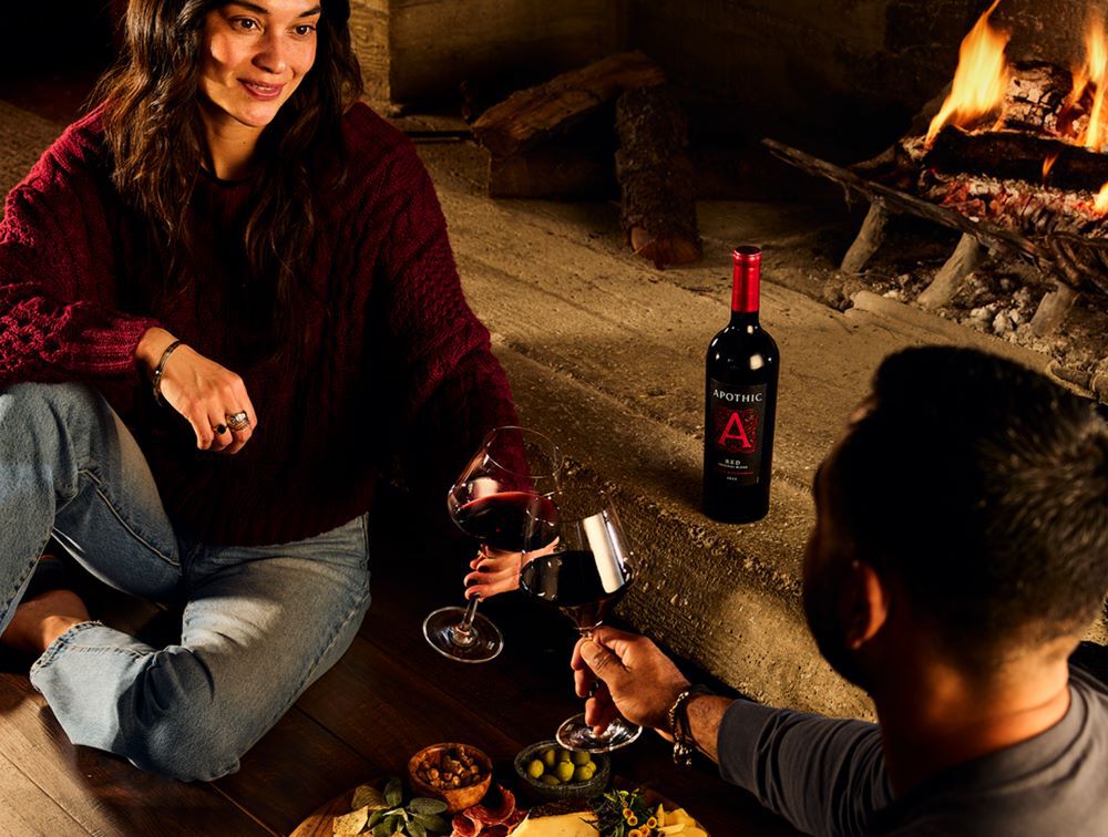 A warm, atmospheric lifestyle shot of a couple enjoying an indoor date night by a glowing fireplace. A woman in a maroon sweater sits on the floor across from her partner, both holding large glasses of red wine. Between them on a low stone ledge is a bottle of Apothic Red Original Blend. In the foreground, a wooden charcuterie board is spread with olives, nuts, and cheeses. The low lighting from the fire creates a cozy, private VIP experience at home.