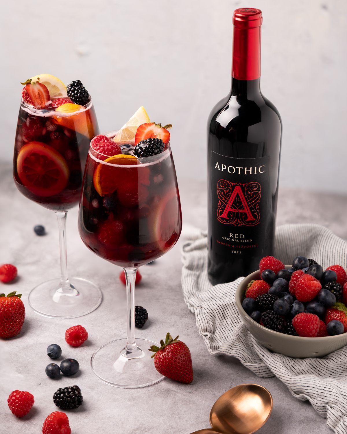 A bottle of Apothic Red 2022 wine stands next to two glasses filled with a dark red wine cocktail garnished with fresh strawberries, lemon slices, and blackberries. A small bowl of mixed berries sits to the side on a striped linen napkin, with loose raspberries, blueberries, and strawberries scattered across a light gray textured surface.
