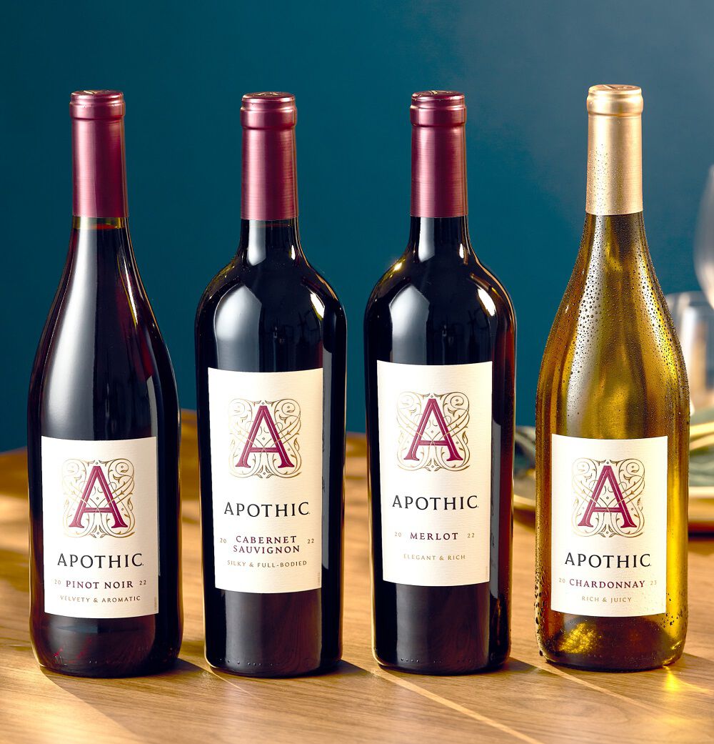 A bright lifestyle shot of four Apothic wine bottles standing on a wooden surface against a teal background. From left to right: Pinot Noir, Cabernet Sauvignon, Merlot, and Chardonnay. The Chardonnay bottle on the far right is chilled with visible condensation. The lighting is crisp and natural, highlighting the white labels and gold and red foil caps.