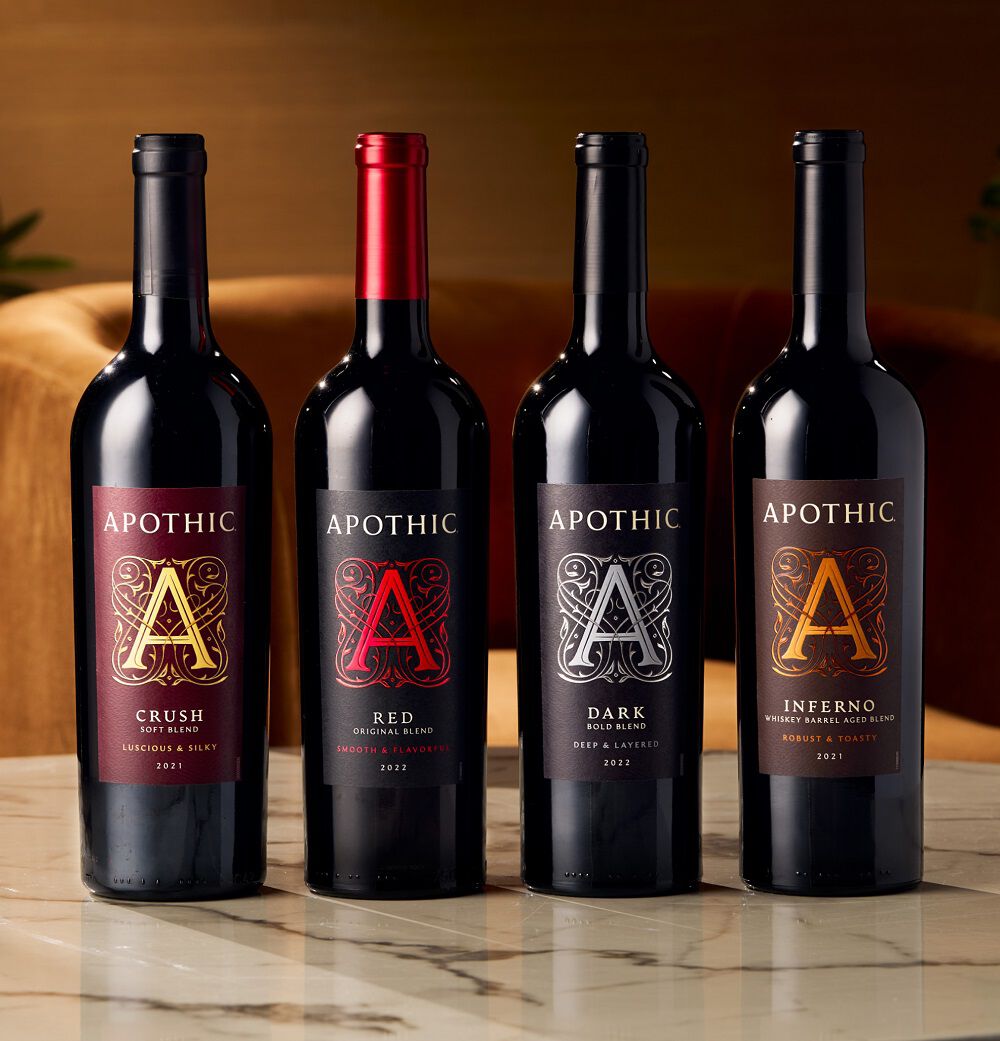 A horizontal lifestyle shot of ten different Apothic wine bottles lined up on a wooden table. From left to right: Dark, White, Inferno, Crush, Red Original Blend, Cabernet Sauvignon, Pinot Noir, Ros&eacute;, Merlot, and Chardonnay. The lighting is warm and moody, with a corkscrew resting on the table in the foreground.