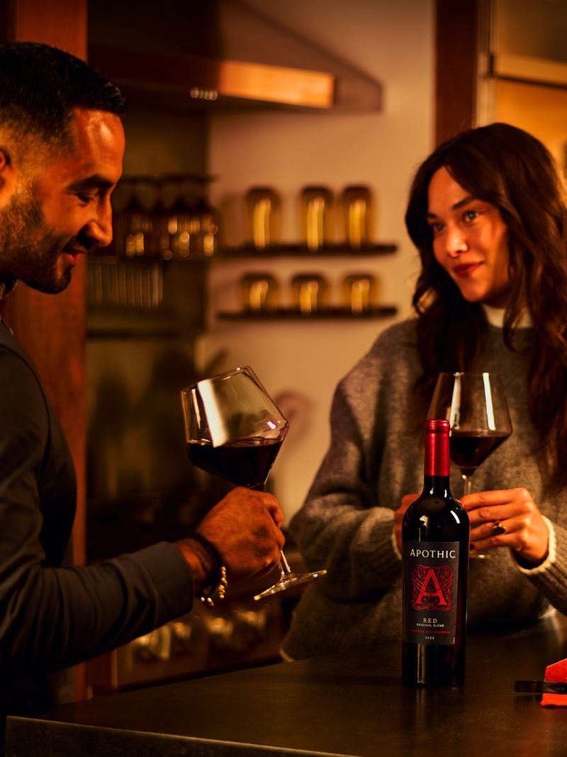 A moody lifestyle photograph of a couple enjoying a private date night in a warmly lit home kitchen. The man on the left and the woman on the right are leaning against a counter, holding glasses of deep red wine and sharing a warm look. In the foreground sits a bottle of Apothic Red Original Blend with its signature large red 'A' logo. The scene feels intimate and crafted for connection, with warm light highlighting their faces and the wine.