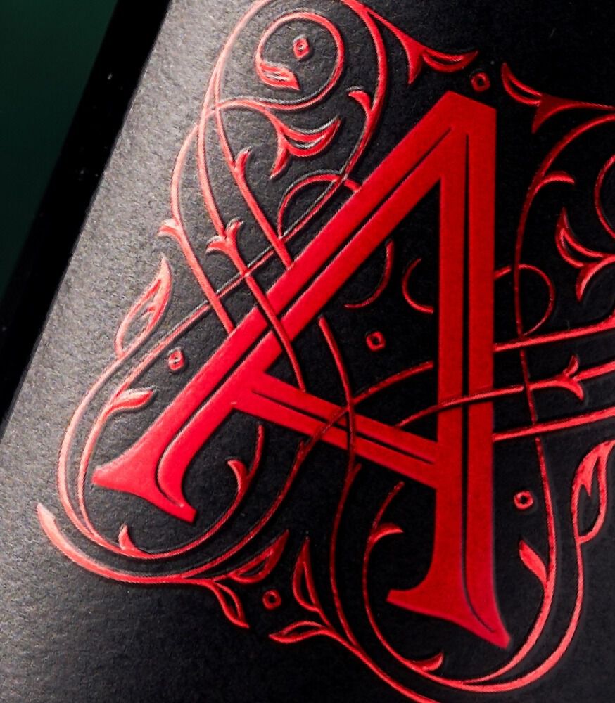 A detailed, macro close-up shot of the Apothic wine logo on a textured black label. A large, stylized capital 'A' is rendered in vibrant, matte red. It is intricately intertwined with detailed, swirling gothic or baroque-style red filigree and vine patterns that are finely debossed into the label paper.
