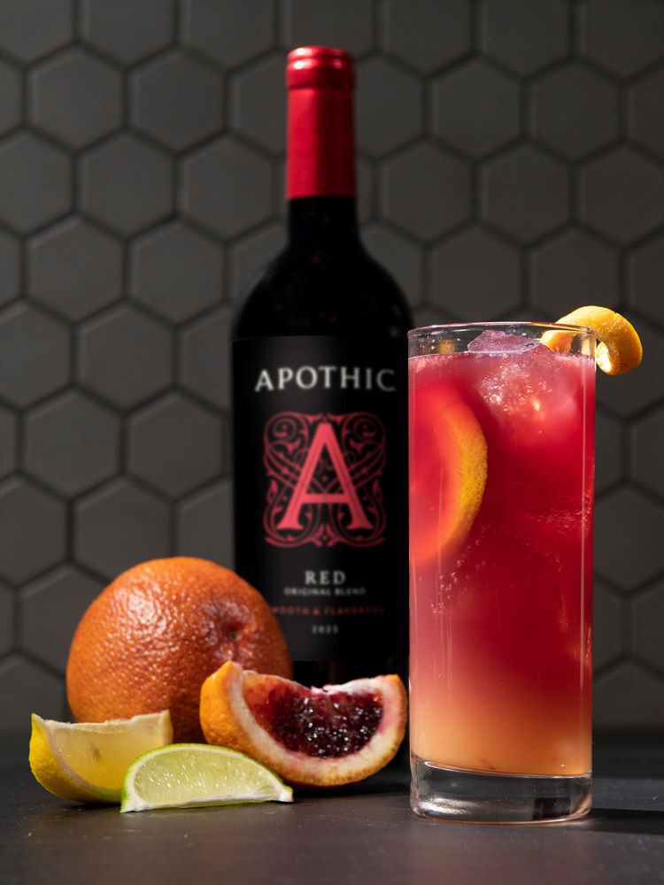 A bright, effervescent Red Sunset cocktail served in a large wine glass on a light, stone-textured surface. The ruby-red drink is filled with ice and topped with a fresh orange slice and a sprig of mint. In the background, a bottle of Apothic Red Original Blend stands next to a small wooden bowl with lemon and lime wedges. The clean, high-key lighting highlights the carbonation and the refreshing citrus garnishes.