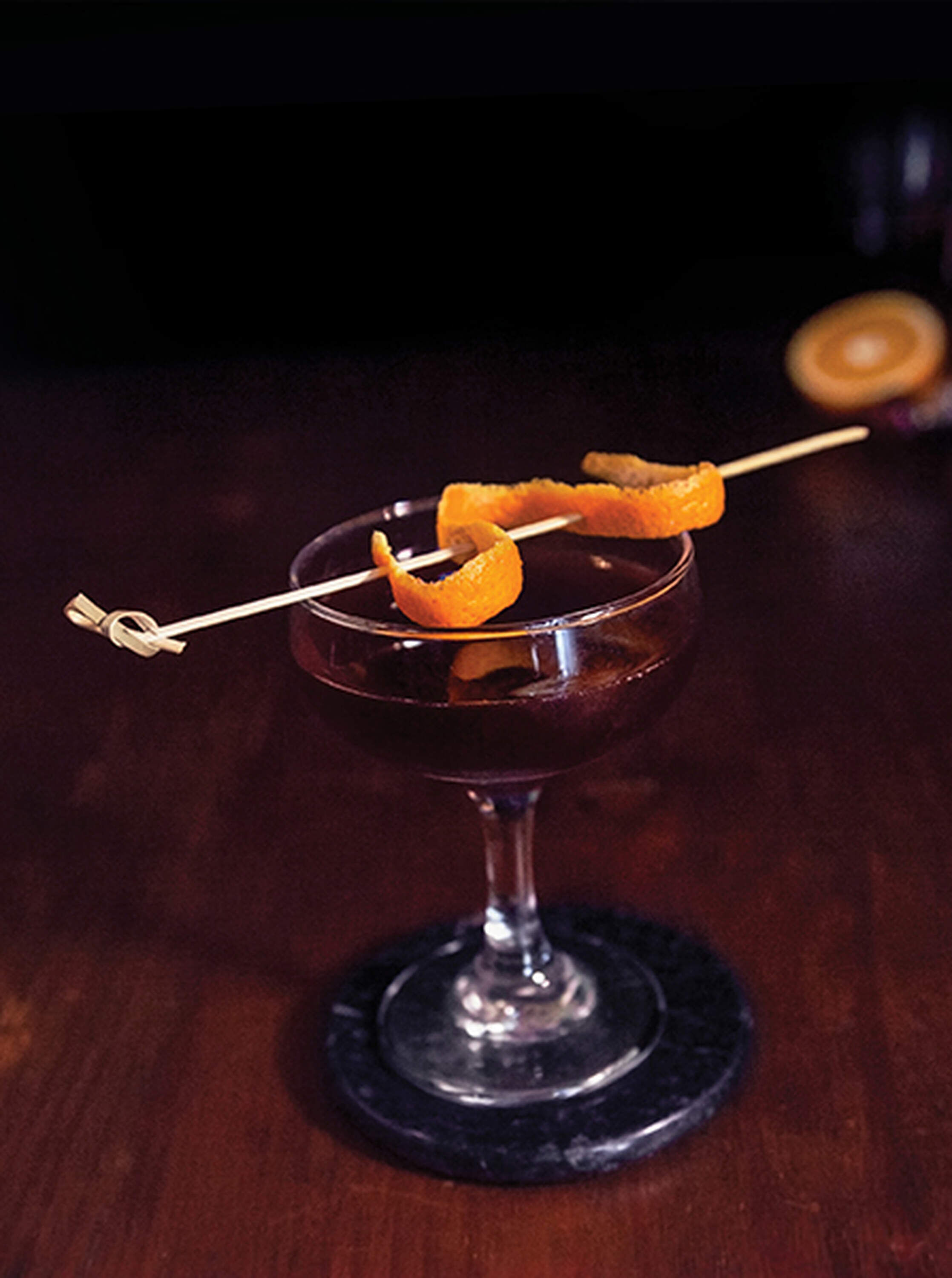 A moody and atmospheric shot of the Firewater cocktail served in a stemmed glass on a dark, reflective surface. The deep red drink is garnished with a single maraschino cherry. In the background, a bottle of Apothic Red Original Blend stands next to a bottle of Scotch whisky and a small bottle of cherry liqueur. Low-intensity lighting creates sharp highlights on the glassware and the liquid, emphasizing the bold, high-contrast aesthetic.