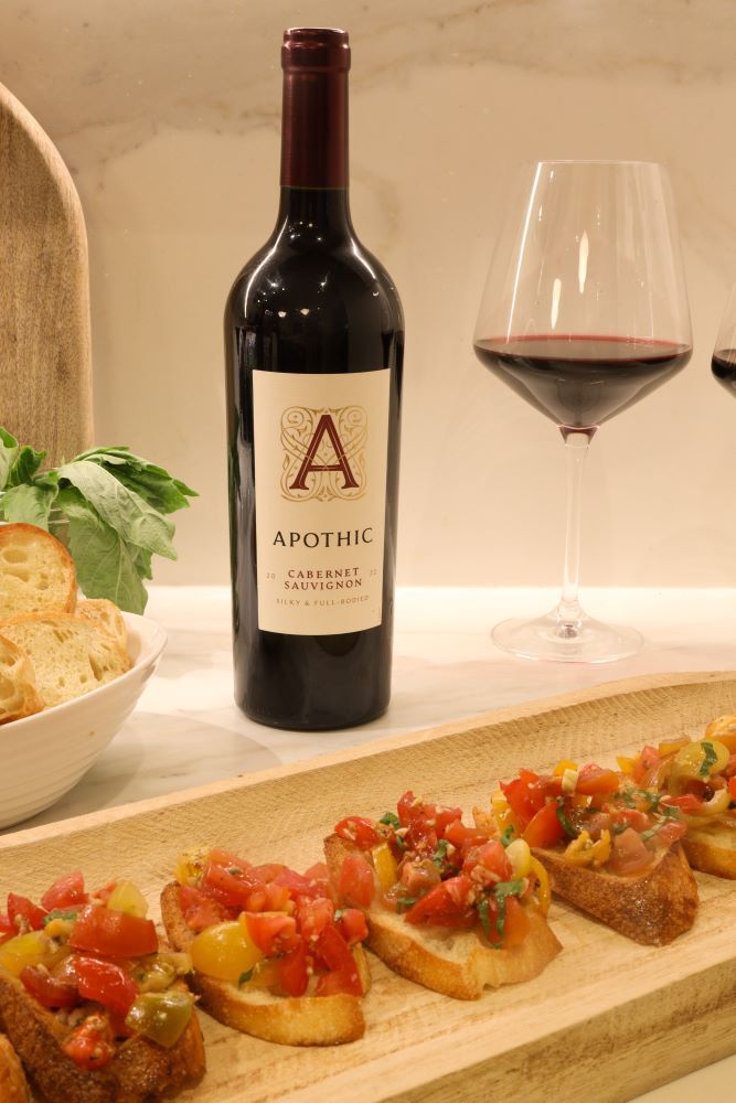 A bright, close-up lifestyle shot of tomato bruschetta appetizers served on a long wooden board. In the background, a bottle of 2022 Apothic Cabernet Sauvignon stands on a white marble countertop next to a glass of red wine. A white bowl of toasted bread and fresh herbs sit to the left, creating a fresh and appetizing food pairing scene.