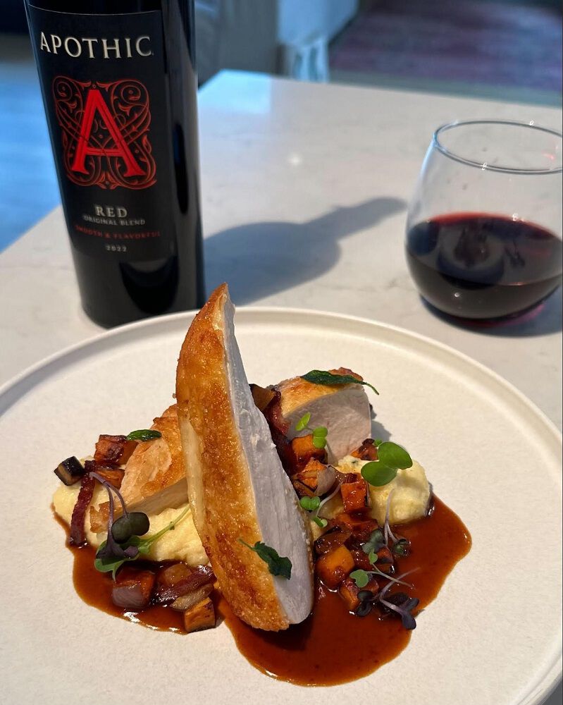 A bright, close-up lifestyle shot of a sliced, pan-seared chicken breast served over a bed of creamy herb polenta and a rich brown sauce, topped with roasted root vegetables and microgreens. In the upper left background, a bottle of 2022 Apothic Red Original Blend stands on a white marble countertop next to a glass of red wine, creating a sophisticated and appetizing dinner scene.