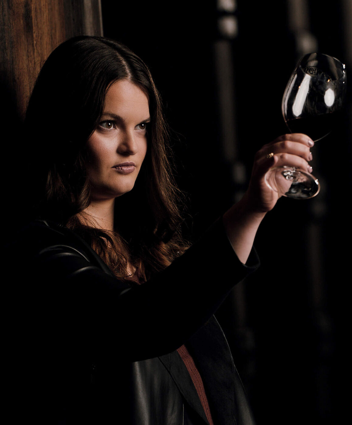 A portrait of Apothic winemaker Ashleigh Ricchio in a dimly lit cellar environment. She is shown from the chest up, wearing a black leather jacket, holding a glass of dark red wine up to the light to examine its color and clarity against a dark, moody background.