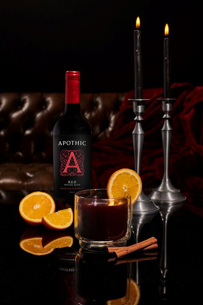 A moody, elegant still life on a reflective black surface, featuring a glass mug of dark red mulled wine garnished with an orange slice. Next to it sit loose orange slices and two cinnamon sticks. Behind the cocktail stands a bottle of Apothic Red Original Blend and two lit silver candlesticks holding black tapers, against a background of dark tufted leather and velvet.