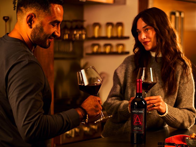 A moody lifestyle photograph of a couple enjoying a private date night in a warmly lit home kitchen. The man on the left and the woman on the right are leaning against a counter, holding glasses of deep red wine and sharing a warm look. In the foreground sits a bottle of Apothic Red Original Blend with its signature large red 'A' logo. The scene feels intimate and crafted for connection, with warm light highlighting their faces and the wine.