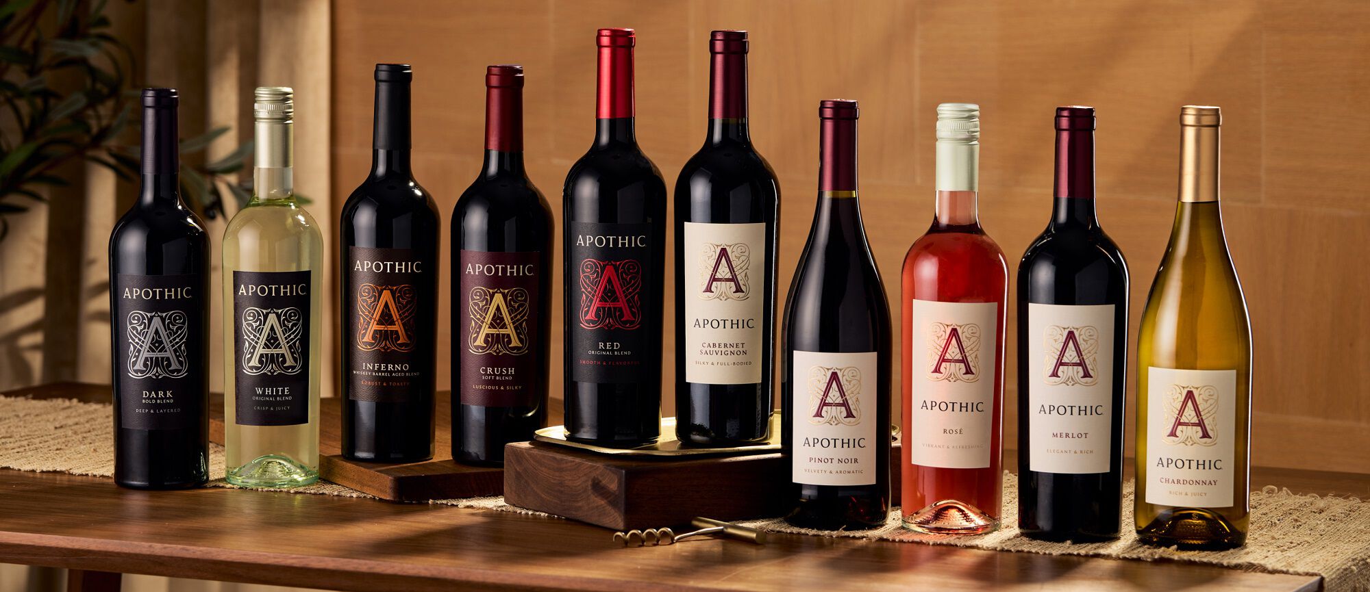 A wide, cinematic shot of ten diverse bottles of Apothic wine arranged in a line on a rustic wooden table. The collection includes Apothic Dark, White, Inferno, Crush, Red, Cabernet Sauvignon, Pinot Noir, Ros&eacute;, Merlot, and Chardonnay. A gold corkscrew sits in the foreground, and the background features warm, soft-focus lighting that highlights the varied colors of the wine from deep reds to pale golds.