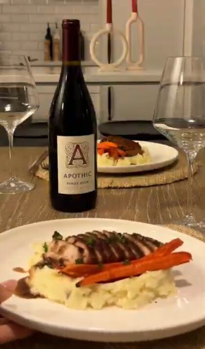 A warm lifestyle photograph of a sliced maple glazed pork chop served over creamy mashed potatoes with roasted carrots on a white plate. In the background, a bottle of Apothic Pinot Noir stands on a wooden dining table between two wine glasses. Another plated serving is visible in the distance, creating a cozy and intimate dinner setting.
