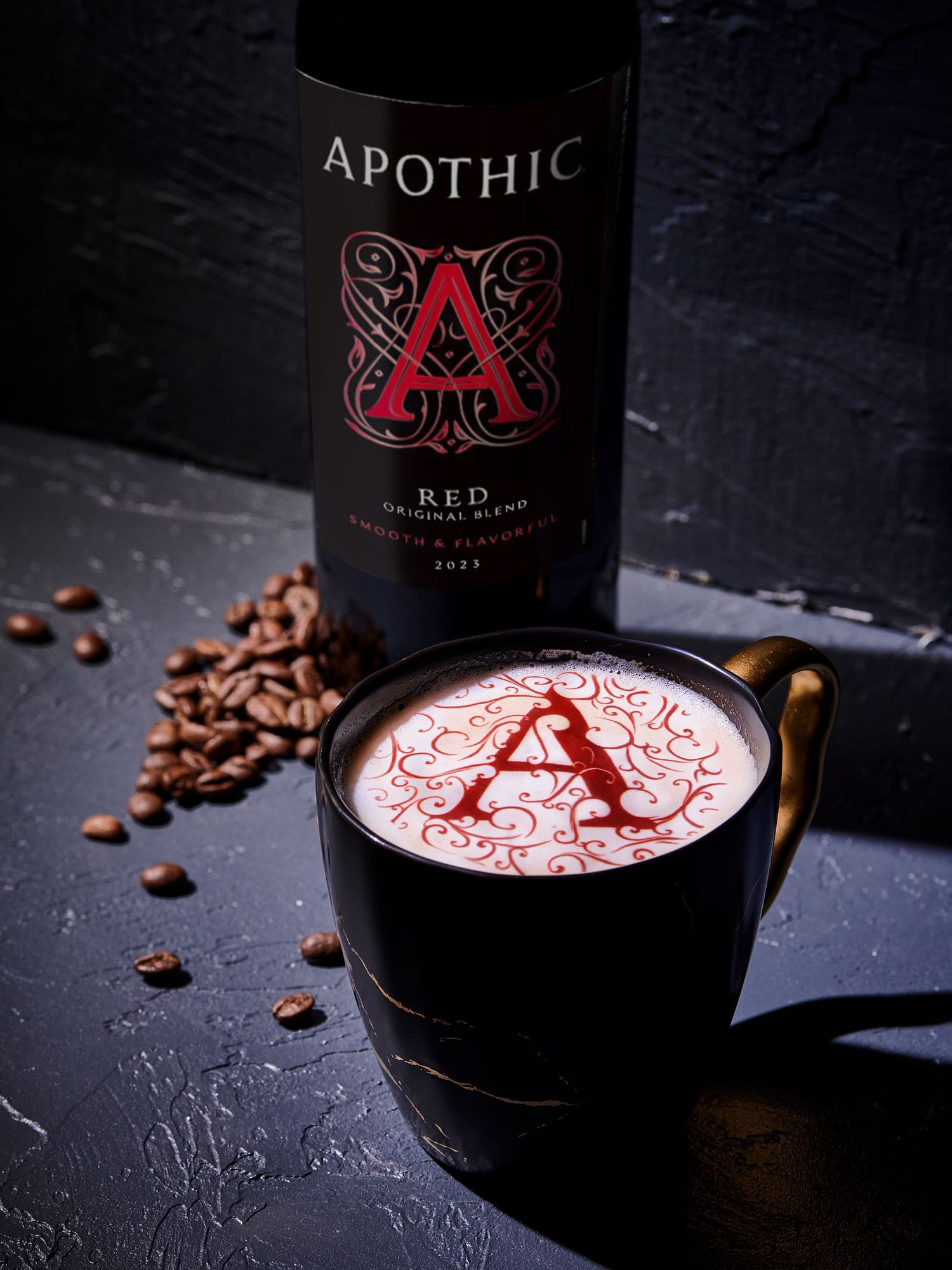 A rich red wine mocha latte served in a clear glass mug on a dark, moody surface. The drink features a thick layer of frothed milk topped with a dusting of cocoa powder and two small dark chocolate squares. To the left, a bottle of Apothic Red Original Blend stands next to a small wooden bowl filled with coffee beans and pieces of dark chocolate. The lighting is warm and atmospheric, highlighting the creamy texture of the foam.