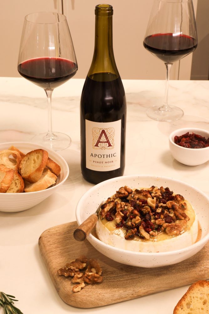 A brightly lit, lifestyle shot of a round of baked brie topped with walnuts and dried cranberries in a white ceramic bowl. The dish sits on a white marble countertop next to a bottle of 2022 Apothic Pinot Noir and two glasses of red wine. To the left, a bowl of sliced baguette and a small wooden board with walnuts complete the sophisticated appetizer scene.