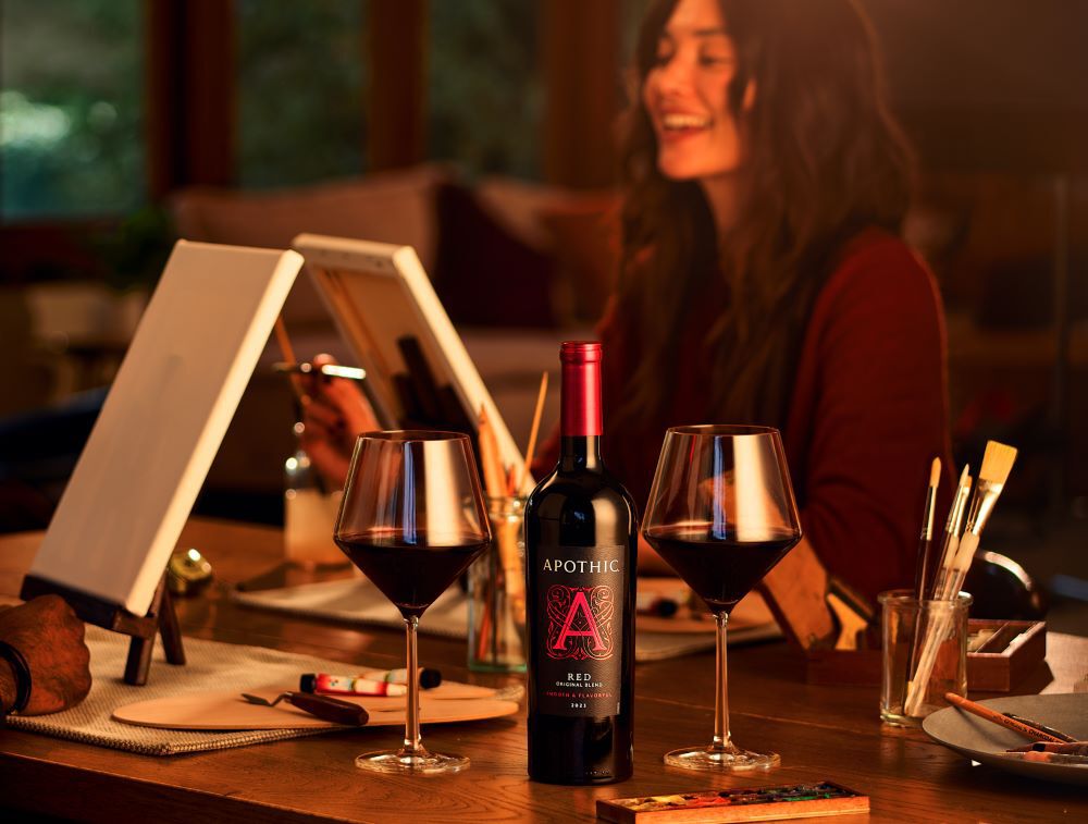 A warm, atmospheric lifestyle photograph of a couple enjoying a creative date night at home. In the center, a bottle of 2023 Apothic Red Original Blend sits between two filled wine glasses on a wooden table. In the blurred background, a woman smiles while painting on a canvas. Art supplies, including brushes and a palette, are scattered across the table, creating a cozy and artistic scene.