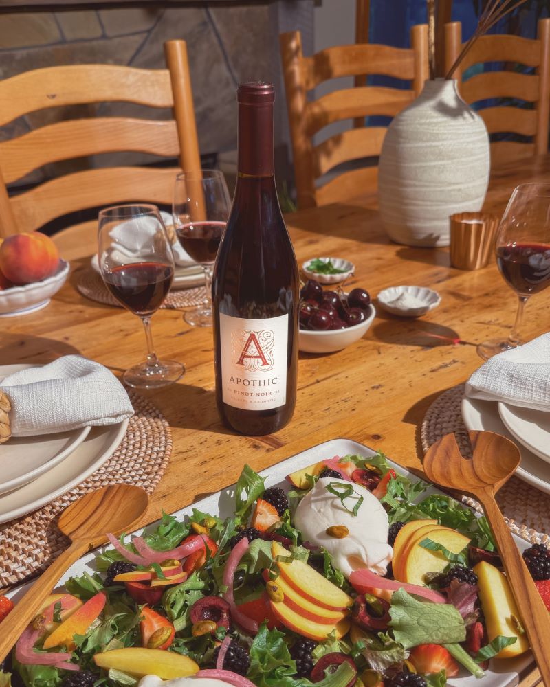 A bright, overhead lifestyle shot of a spring peach and burrata salad served in a shallow white ceramic bowl. The dish features large pieces of creamy white burrata cheese, sliced yellow peaches, fresh basil leaves, and a drizzle of balsamic glaze. A bottle of 2023 Apothic Chardonnay and a glass of white wine are positioned at the top of the frame on a light-colored tabletop, surrounded by fresh peach slices and basil sprigs.