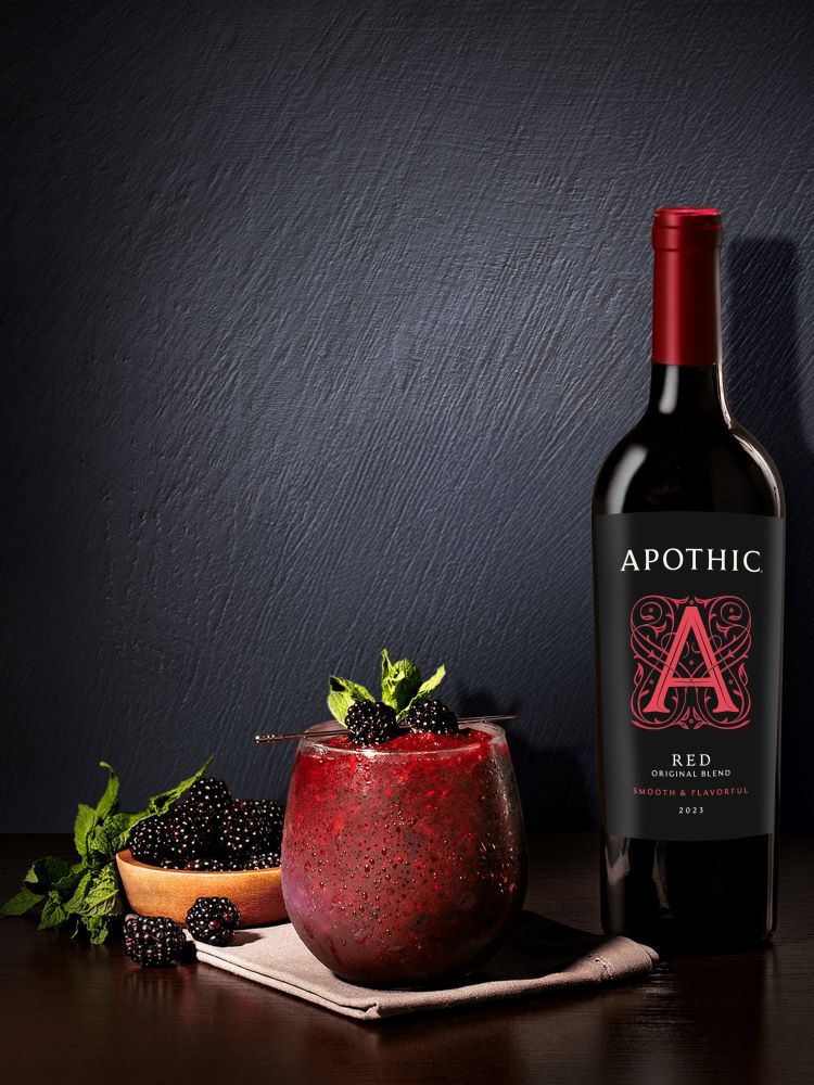 A moody, high-contrast photograph of a Black Ice cocktail on a dark textured surface. A stemless glass is filled with a deep purple, crushed-ice wine blend, garnished with two fresh blackberries and a sprig of mint on a cocktail pick. To the right, a bottle of 2023 Apothic Red Original Blend stands against a dark gray wall. To the left, a small wooden bowl overflows with fresh blackberries and mint leaves.