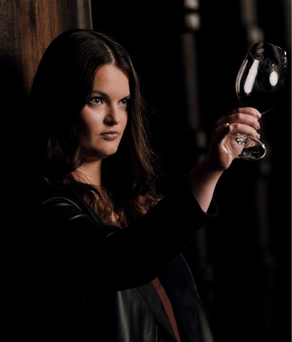 A portrait of Apothic winemaker Ashleigh Ricchio in a dimly lit cellar environment. She is shown from the chest up, wearing a black leather jacket, holding a glass of dark red wine up to the light to examine its color and clarity against a dark, moody background.