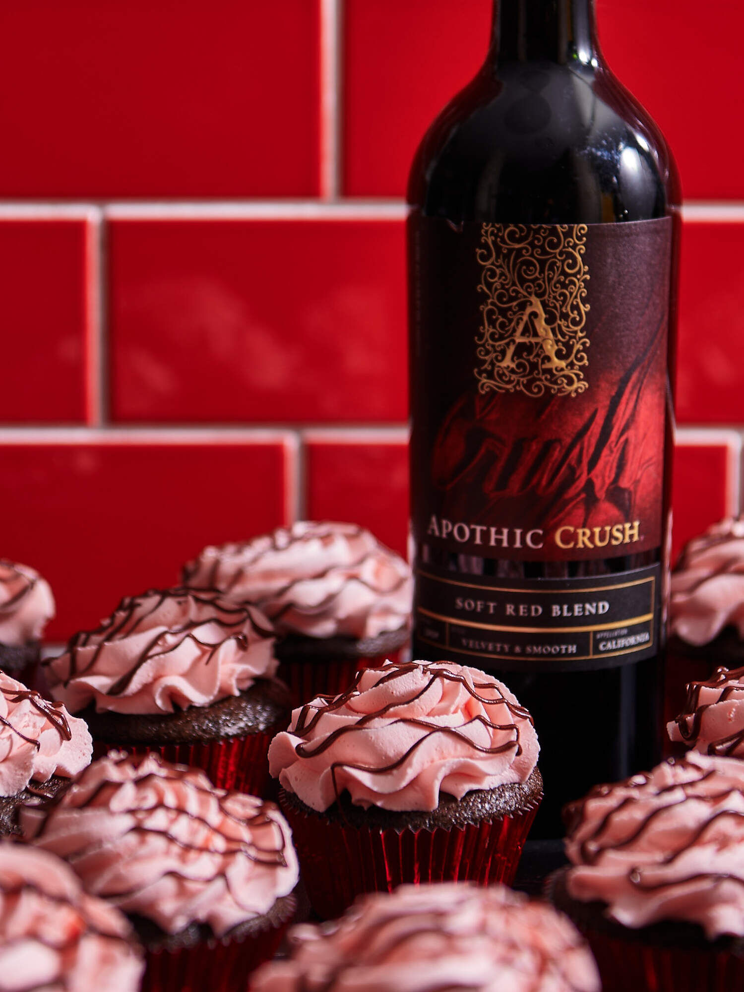 A bright, close-up lifestyle photograph of several chocolate cupcakes with swirls of light pink frosting drizzled with dark chocolate. In the background, a bottle of Apothic Crush Soft Red Blend stands against a vibrant red tiled wall. The cupcakes are arranged in a group, highlighting their velvety texture and the 'Velvety & Smooth' profile of the wine pairing.