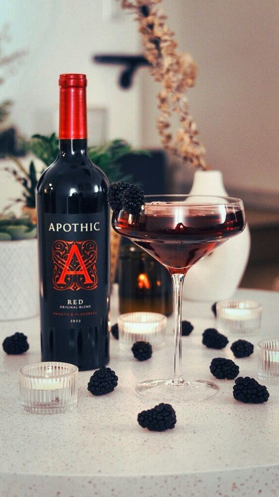 A sophisticated lifestyle shot of a Red Martini served in a chilled martini glass on a dark, reflective surface. The deep crimson drink is garnished with a single maraschino cherry. In the background, a bottle of Apothic Red Original Blend stands next to a clear glass bottle of gin and a small glass of simple syrup. Warm, focused lighting highlights the condensation on the glass and the rich color of the cocktail blend.