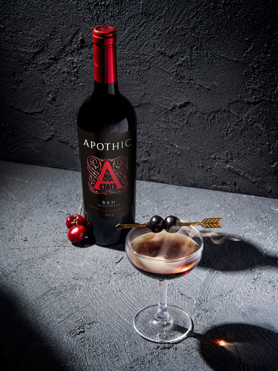 A bottle of Apothic Red 2022 wine stands next to two glasses filled with a dark red wine cocktail garnished with fresh strawberries, lemon slices, and blackberries. A small bowl of mixed berries sits to the side on a striped linen napkin, with loose raspberries, blueberries, and strawberries scattered across a light gray textured surface.
