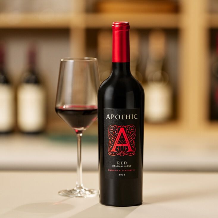 A bottle of Apothic Red Original Blend 2022 wine stands next to a partially filled wine glass on a light-colored counter. The bottle features a black label with a large, ornate red 'A'. The background is a soft-focus view of a warm, wooden shelf with other wine bottles.