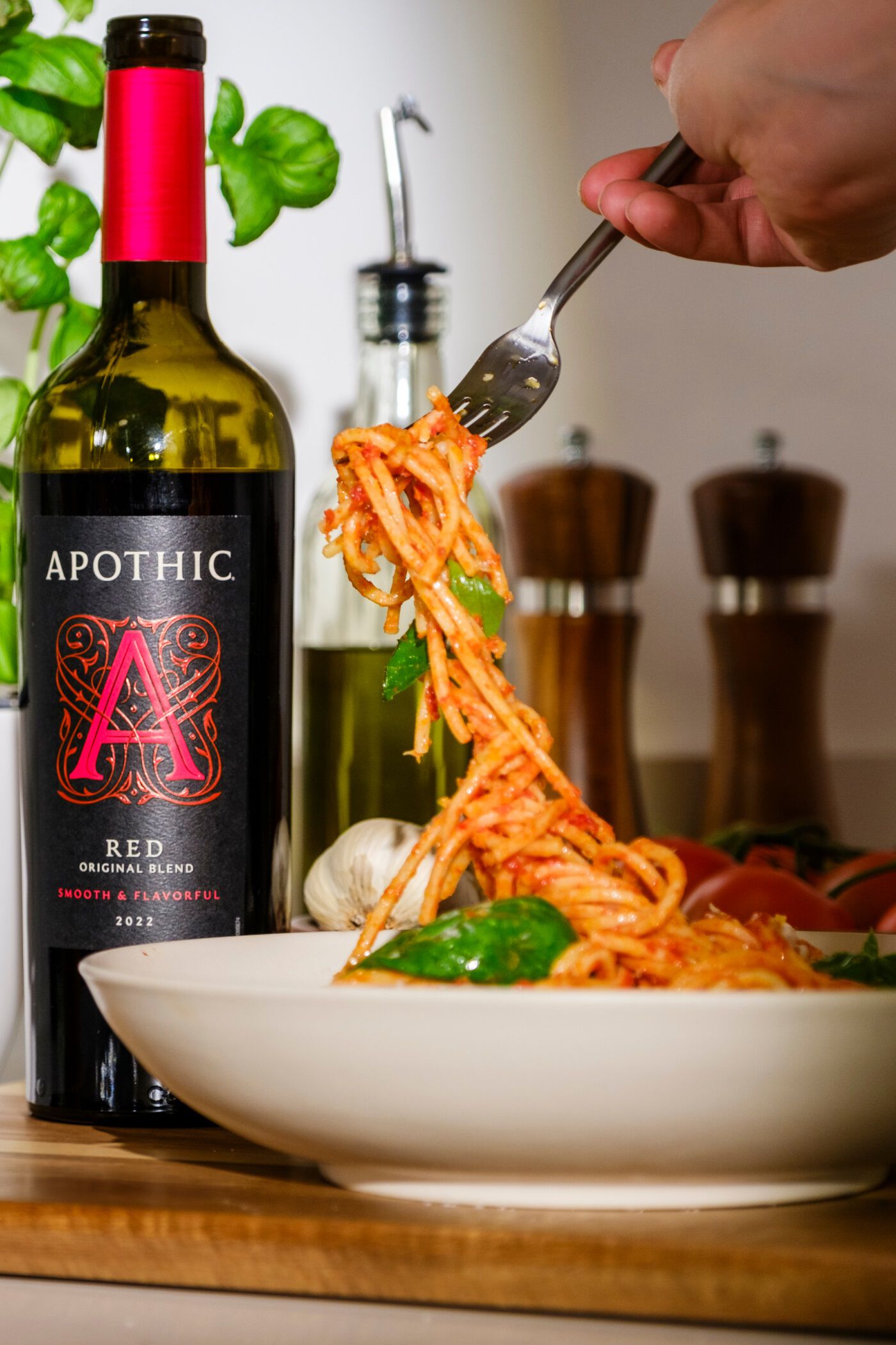 A close-up, dynamic lifestyle shot of a fork twirling spaghetti coated in red pomodoro sauce and fresh basil over a white bowl. To the left, a bottle of 2022 Apothic Red Original Blend stands prominently. The background features a rustic kitchen setting with fresh basil, a head of garlic, tomatoes, and wooden salt and pepper grinders on a wooden countertop.