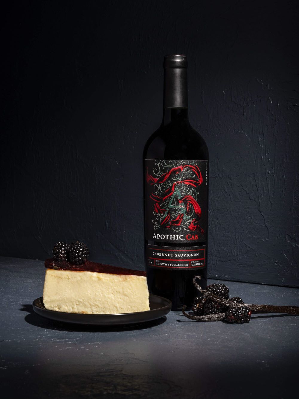 A sophisticated, dark-toned lifestyle photograph featuring a slice of creamy vanilla cheesecake topped with a dark berry glaze and two fresh blackberries. In the background, a bottle of Apothic Cab Cabernet Sauvignon stands on a textured dark gray surface, accompanied by fresh blackberries and vanilla beans to emphasize the wine's flavor profile.