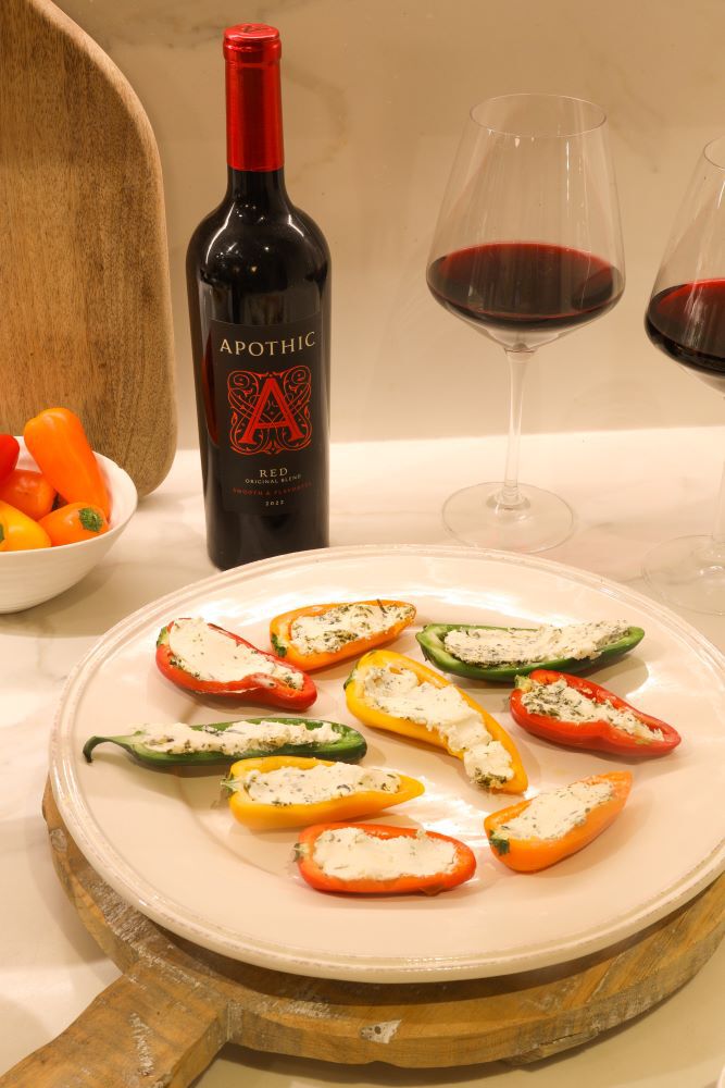 A bright lifestyle photograph of a white plate with eight colorful mini peppers&mdash;red, orange, and green&mdash;halved and stuffed with a creamy herb cheese spread. In the background, a bottle of 2022 Apothic Red Original Blend stands on a white marble countertop next to two filled wine glasses. A white bowl of whole mini peppers and a wooden cutting board are visible on the left, creating a vibrant and fresh appetizer setting.