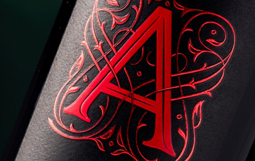 A detailed, macro close-up shot of the Apothic wine logo on a textured black label. A large, stylized capital 'A' is rendered in vibrant, matte red. It is intricately intertwined with detailed, swirling gothic or baroque-style red filigree and vine patterns that are finely debossed into the label paper.