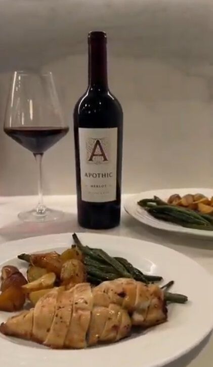 A warm lifestyle photograph featuring a sliced honey garlic chicken breast served on a white plate with roasted potatoes and green beans. In the background, a bottle of Apothic Merlot and a glass of red wine are positioned on a white countertop, creating a cozy and complete dinner setting.