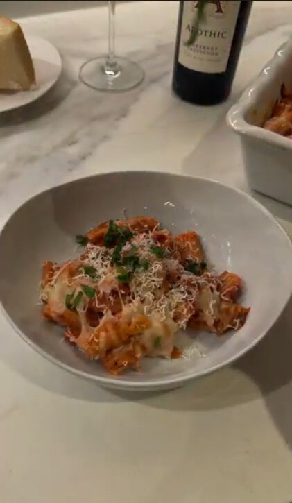 A warm, overhead lifestyle shot of a white rectangular baking dish filled with classic baked ziti, featuring melted mozzarella cheese, tomato sauce, and fresh basil leaves. In the background, a bottle of 2022 Apothic Red Original Blend and a glass of red wine are set on a white marble countertop alongside a small bowl of fresh basil and a wooden spoon.