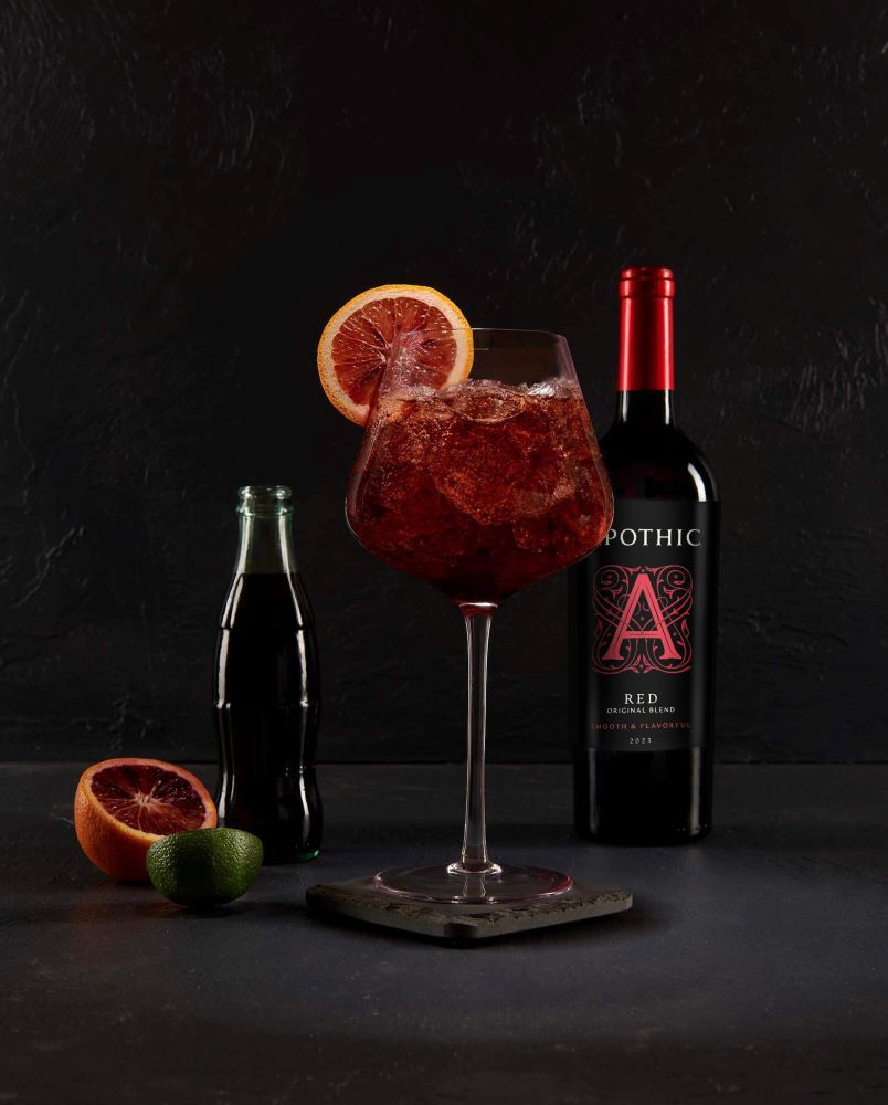 A refreshing Kalimotxo cocktail served in a tall fluted glass on a light stone surface. The drink is a deep, dark red with visible carbonation bubbles and is garnished with a fresh lemon wheel. In the background, a bottle of Apothic Red Original Blend stands next to a small wooden bowl containing lemon and lime wedges. The lighting is bright and clean, highlighting the effervescence of the cola blend.
