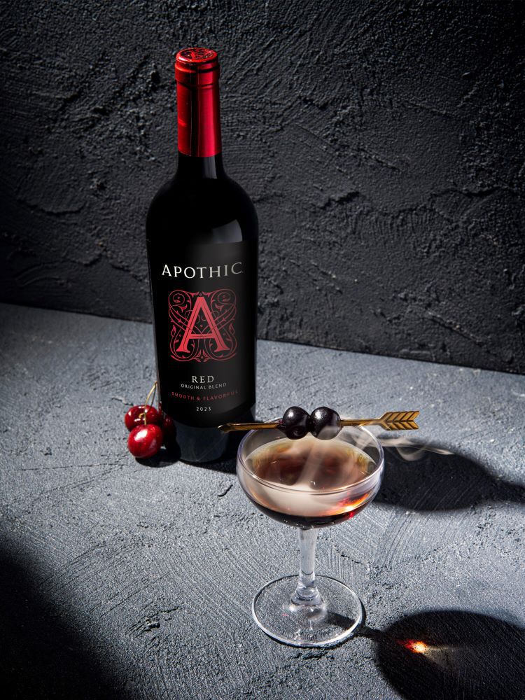 A moody, close-up lifestyle photograph of the Red Passion cocktail, featuring a deep red liquid in a faceted crystal rocks glass. The drink is garnished with a single maraschino cherry on a cocktail pick and a long, thin orange peel twist. In the background, a bottle of Apothic Red Original Blend and a crystal decanter of bourbon sit on a dark, textured surface under warm, focused lighting.