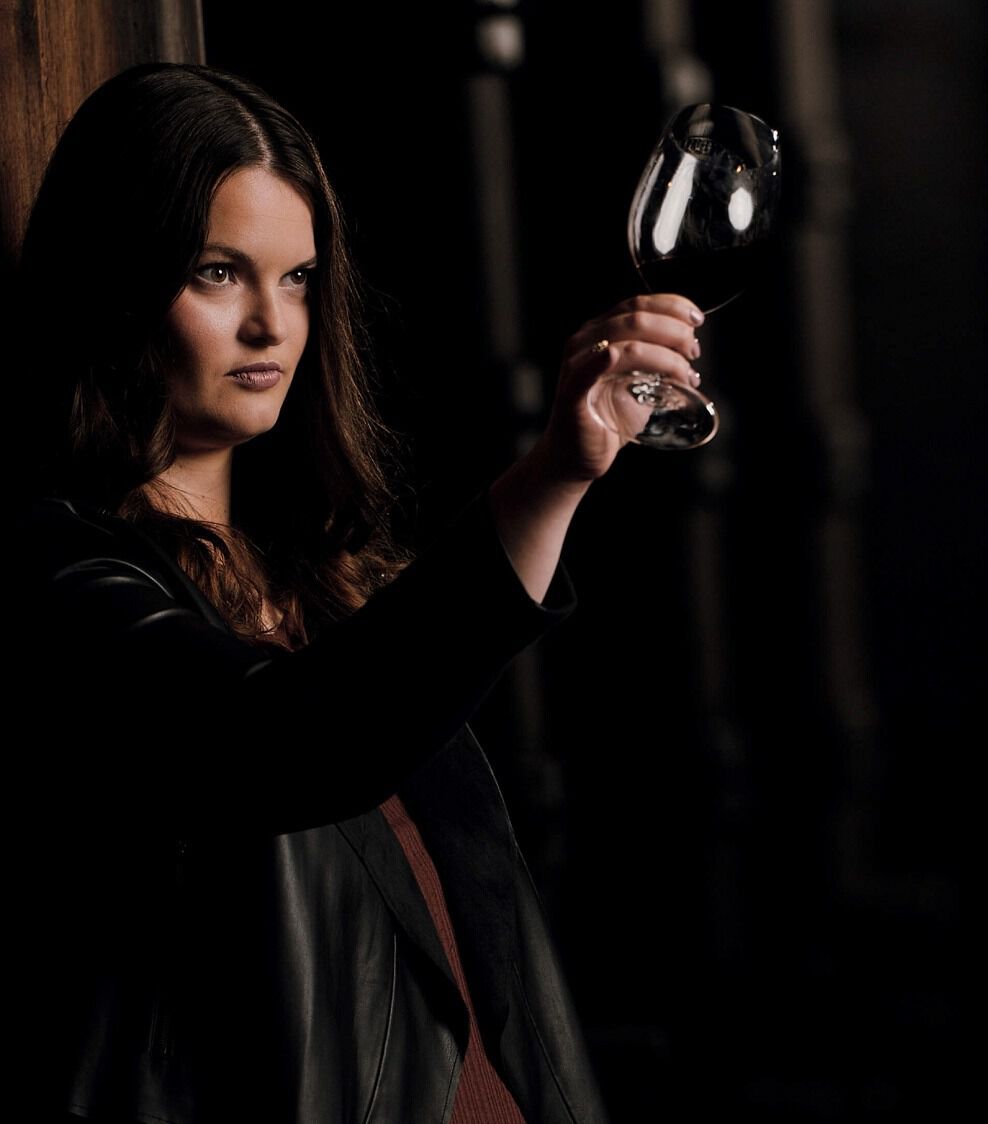 A moody, low-light portrait of Apothic winemaker Ashleigh 'Ash' Ricchio. She is leaning against a dark wooden surface, wearing a black leather jacket, and looking thoughtfully off-camera. She holds a glass of deep red wine up to the light, which catches the rim of the glass and highlights her face against the dark, shadowed background.