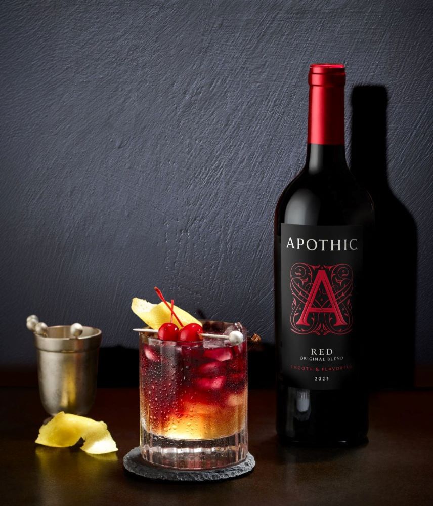 A sophisticated Red Sour cocktail served in a crystal rocks glass on a dark, reflective surface. The drink features a deep red base with a thick, frothy white foam head, garnished with a single maraschino cherry. To the left, a bottle of Apothic Red Original Blend stands next to a crystal decanter of bourbon and a small bowl of fresh lemons. The lighting is moody and focused, casting elegant shadows and highlighting the creamy texture of the foam.